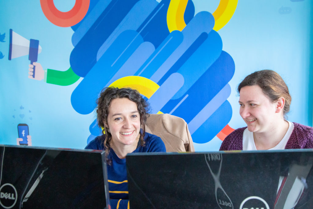 Deux collaboratrices MV Group souriant, derrière leurs écrans, mur bleu en fond