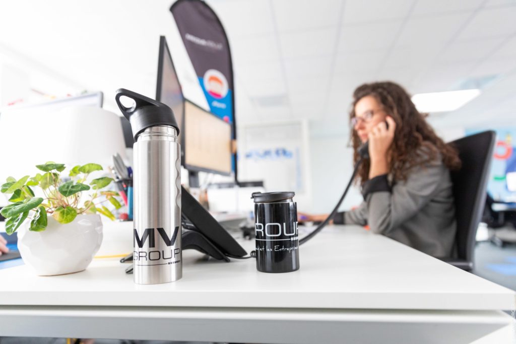 Premier plan : gourdes MV Group sur un bureau - Collaboratrice MV Group au téléphone en arrière-plan